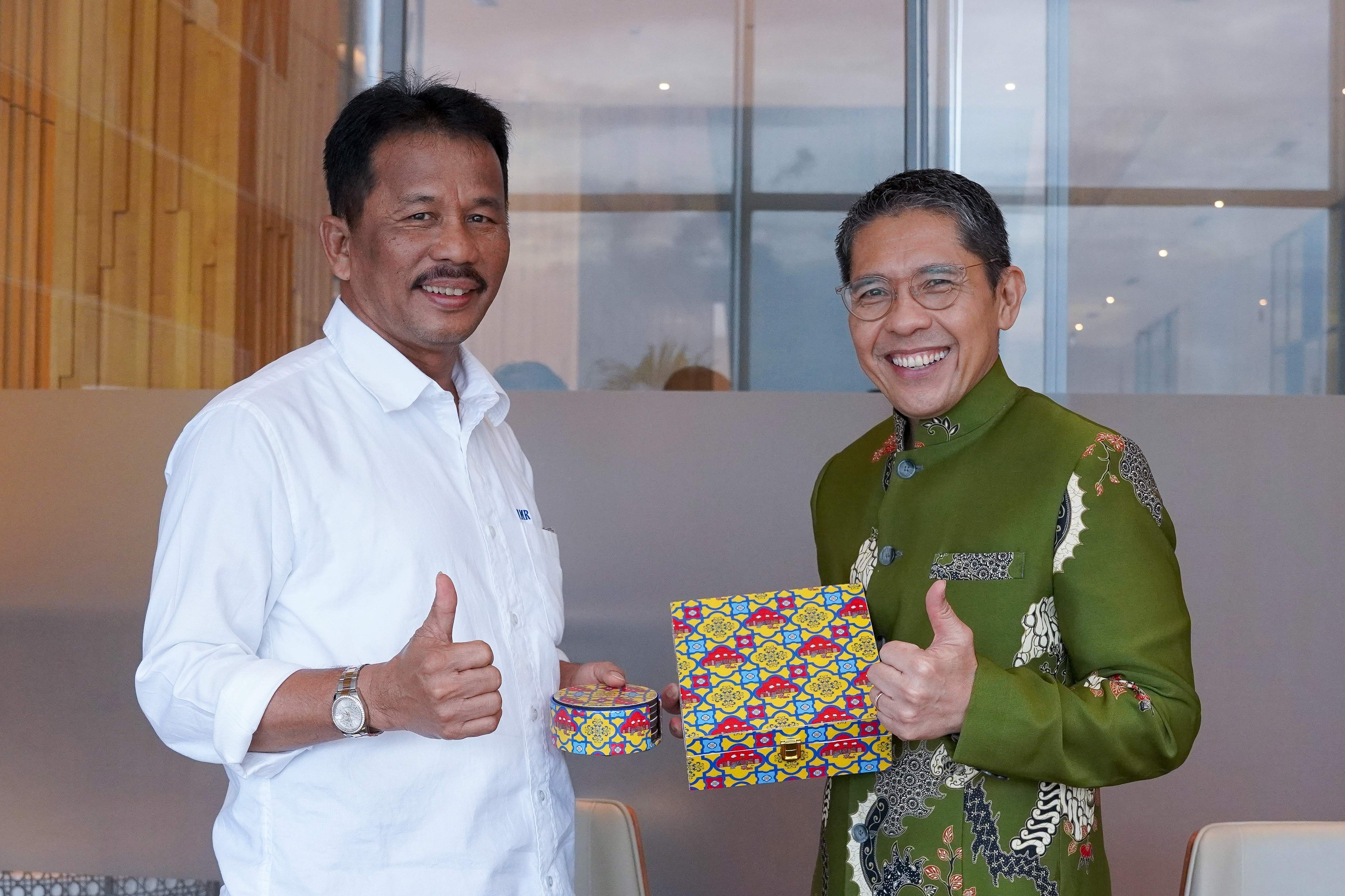 Two men hold patterned boxes, give thumbs up. One in a white shirt, the other in a green batik jacket.